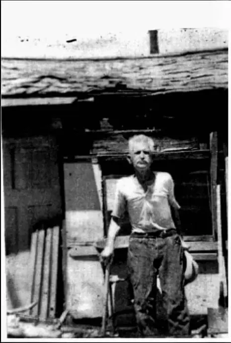 Un retrato fotográfico antiguo en blanco y negro que muestra a William Henry "Burro" Schmidt de pie frente a una estructura rústica.

Persona: Un hombre de edad avanzada con cabello blanco y bigote, vestido con una camisa clara de manga corta y pantalones oscuros desgastados. Sostiene lo que parece ser una herramienta o bastón con su mano derecha.

Entorno: Se encuentra de pie en el exterior, justo delante de una edificación de madera vieja, precaria o de aspecto minero. En el suelo y a su alrededor se aprecian maderas, herramientas y escombros propios de una zona de trabajo rústica.

Estilo: La imagen tiene una calidad granulada, de alto contraste y envejecida, característica de los registros fotográficos de mediados del siglo XX.