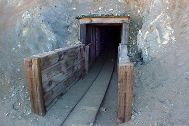 Una fotografía a color de la entrada al túnel de William Henry "Burro" Schmidt, ubicado en una ladera árida y desértica.

La entrada: La boca del túnel está excavada directamente en la roca clara y tierra de la colina. Cuenta con una estructura de soporte rústica de madera vieja y metal corrugado que enmarca la entrada, abriéndose hacia un interior completamente oscuro.

El entorno: El túnel está situado en una pendiente empinada de terreno arenoso y rocoso de color beige o gris claro, salpicada por arbustos secos y vegetación baja desértica. El cielo arriba es de un azul despejado.