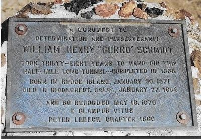 Una fotografía de una placa conmemorativa de metal rectangular, sujeta con grandes tornillos sobre una base de piedra o cemento.

Texto de la placa: Contiene una inscripción grabada en inglés con letras mayúsculas que rinde homenaje a William Henry "Burro" Schmidt. El texto destaca su determinación y perseverancia al cavar a mano un túnel de media milla de largo durante 38 años, completado en 1938. También incluye sus fechas de nacimiento (30 de enero de 1871 en Rhode Island) y fallecimiento (27 de enero de 1954 en Ridgecrest, California), así como la fecha de registro del monumento (16 de mayo de 1970) por la organización E Clampus Vitus, capítulo Peter Lebeck 1866.

Apariencia: La placa de bronce o hierro tiene una pátina envejecida con tonos grisáceos y azulados debido a la exposición a la intemperie.