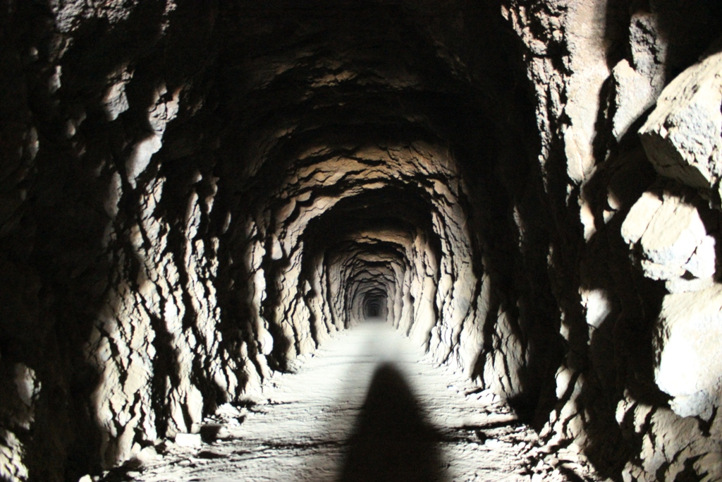 Una fotografía a color tomada desde el interior del túnel de William Henry "Burro" Schmidt, mirando hacia la profundidad del pasaje.

Estructura del túnel: Las paredes y el techo muestran una superficie rugosa e irregular de roca sólida tallada a mano, revelando las marcas del trabajo de excavación. El túnel es estrecho, de forma arqueada y se extiende en línea recta hacia el fondo.

Iluminación: La escena está iluminada artificialmente, posiblemente por la linterna o el flash del fotógrafo, lo que genera fuertes contrastes entre las zonas iluminadas del suelo y los relieves rocosos, y las sombras oscuras en los recovecos de las paredes.

Suelo y perspectiva: El suelo es de tierra o roca aplanada y polvorienta, mostrando en primer plano la sombra alargada de quien toma la fotografía. La perspectiva crea un efecto de profundidad que dirige la mirada hacia el punto más lejano y oscuro del túnel.