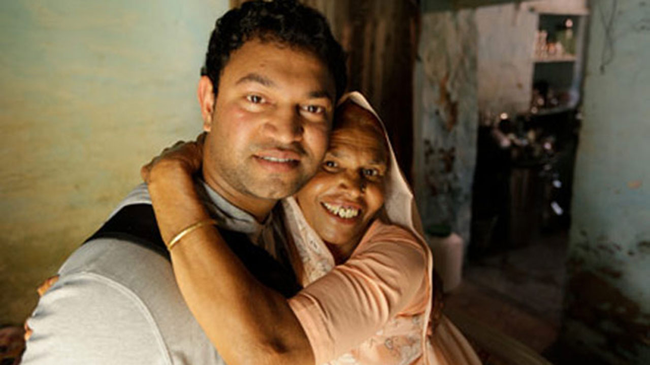 Una fotografía a color de plano medio que muestra a Saroo Brierley (nacido como Sheru Munshi Khan) abrazando afectuosamente a su madre biológica, Kamla Munshi, tras su reencuentro.

Saroo Brierley (Izquierda): Un hombre joven con el cabello corto y oscuro, que viste una camiseta gris claro. Mira directamente a la cámara con una sonrisa leve y serena mientras sostiene a su madre.

Kamla Munshi (Derecha): Una mujer con un velo o bindi tradicional claro sobre la cabeza y vistiendo una prenda de color salmón o rosa pálido. Abraza a su hijo pasando su brazo por el cuello de él y sonríe con gran emoción y alegría hacia la cámara, mostrando los dientes.

Entorno: Se encuentran en el interior de una vivienda humilde con paredes desgastadas de tono azul claro o verdoso. Al fondo, a la derecha, se observa una apertura que da hacia otra habitación más oscura de la casa.