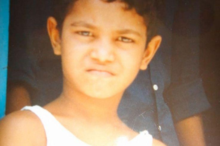 Un primer plano fotográfico a color de un niño pequeño, identificado como Saroo Brierley en su infancia.

Rostro y expresión: El niño mira fijamente hacia la cámara con una expresión seria y el ceño ligeramente fruncido. Tiene el cabello oscuro y corto.

Atuendo: Lleva una camiseta blanca de tirantes.

Fondo: Detrás de él se observa de manera difuminada el torso de otra persona vestida con una camisa oscura, y a la izquierda se aprecia un fragmento de una superficie o pared de color azul brillante.